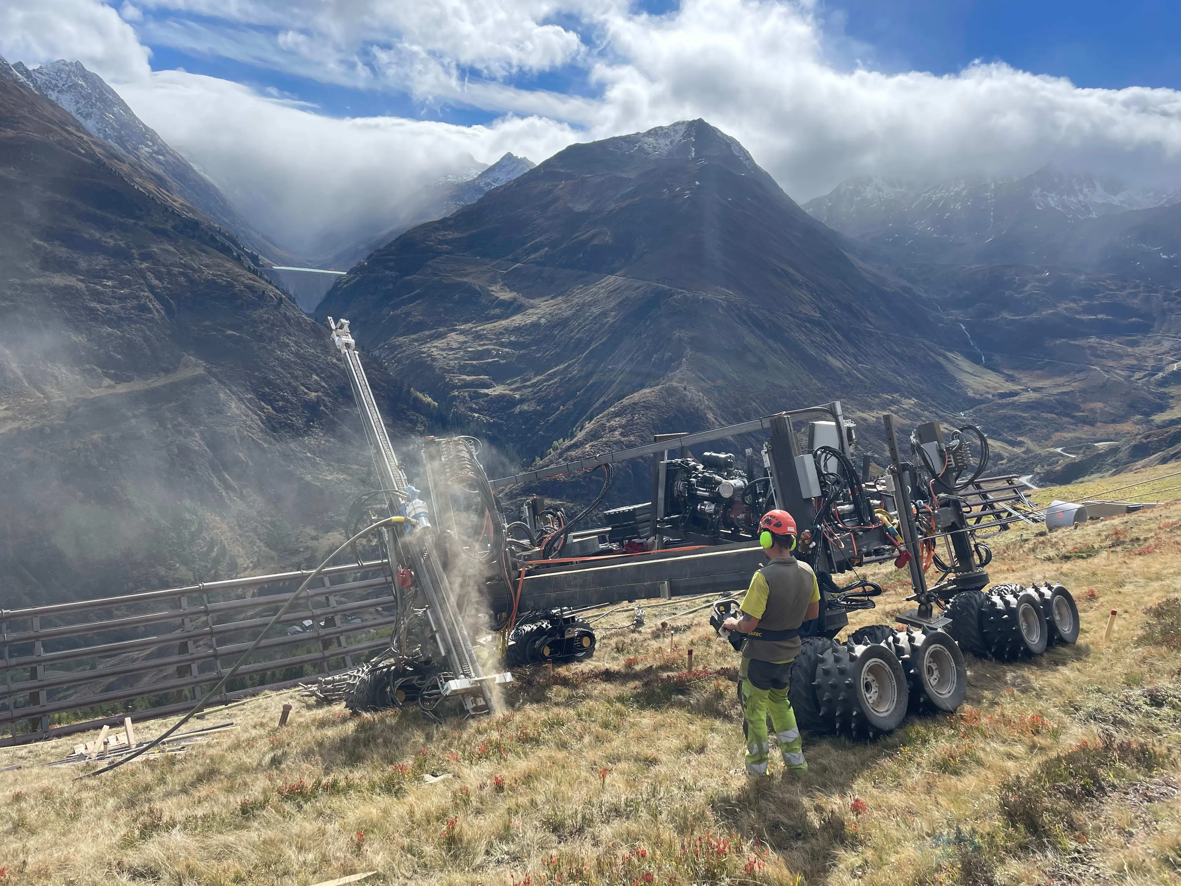 A worker in a red helmet and high-vis gear operates heavy drilling machinery on a mountain slope with alpine scenery.