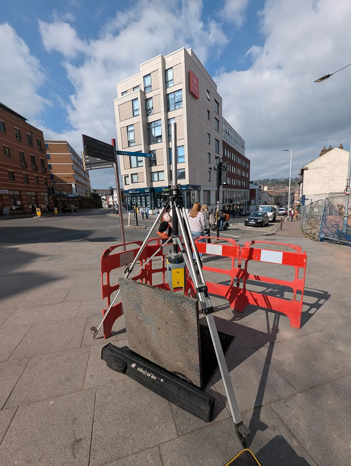 A Trimble surveying tripod set up on a city sidewalk, surrounded by red barriers, with a modern building and busy street in the background.