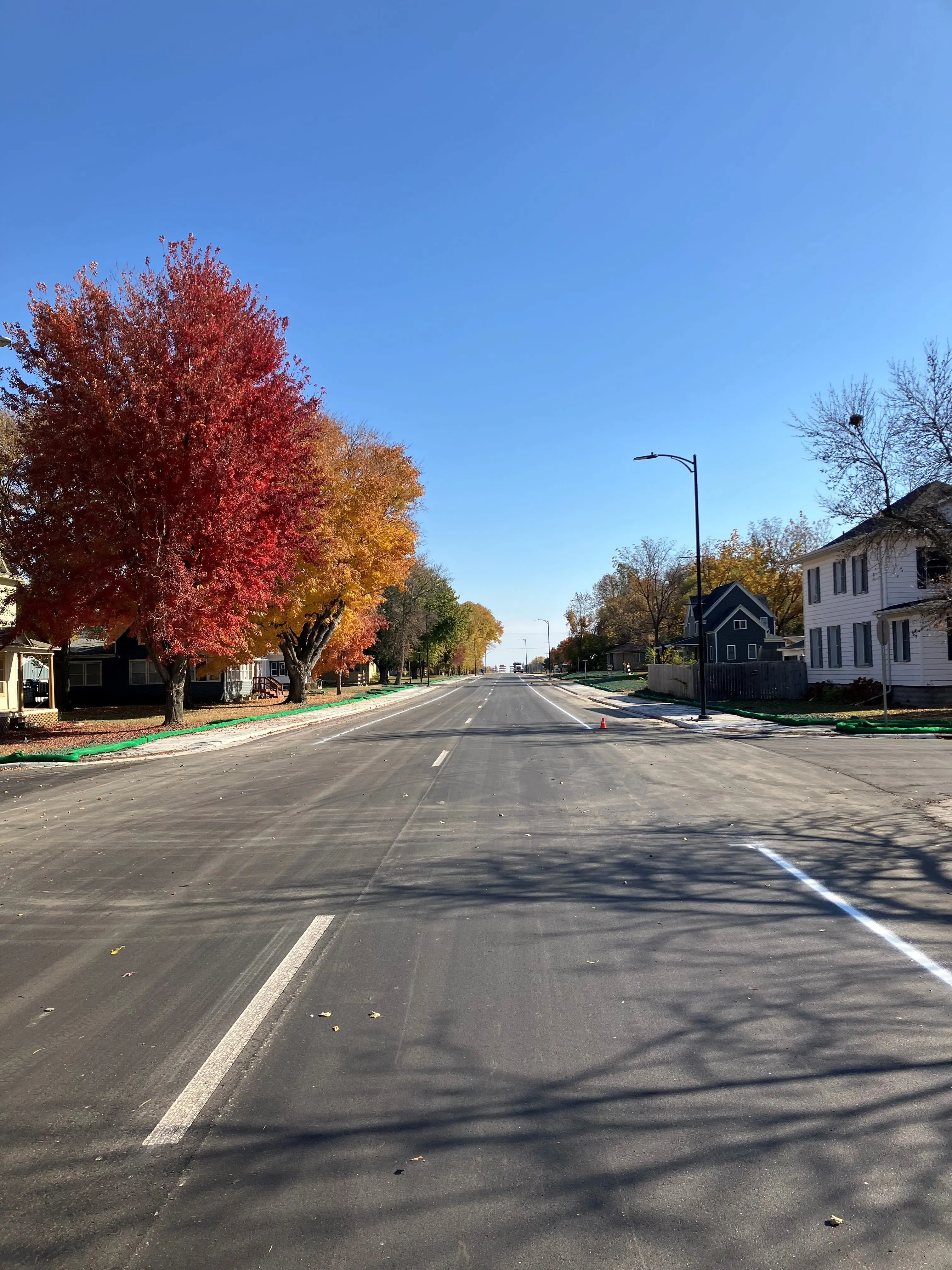A quiet residential street in autumn, lined with vibrant red and orange trees, houses, and a clear blue sky above.