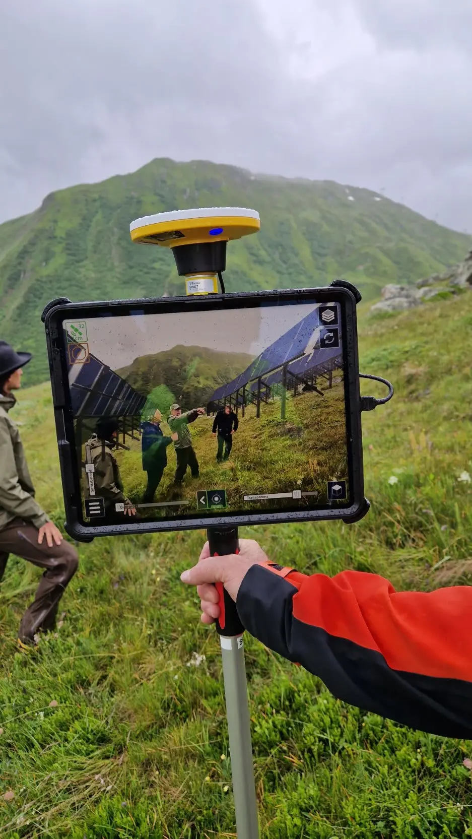 A person holds a Trimble GPS-equipped tablet outdoors, displaying an AR overlay of solar panels on a green mountainside.