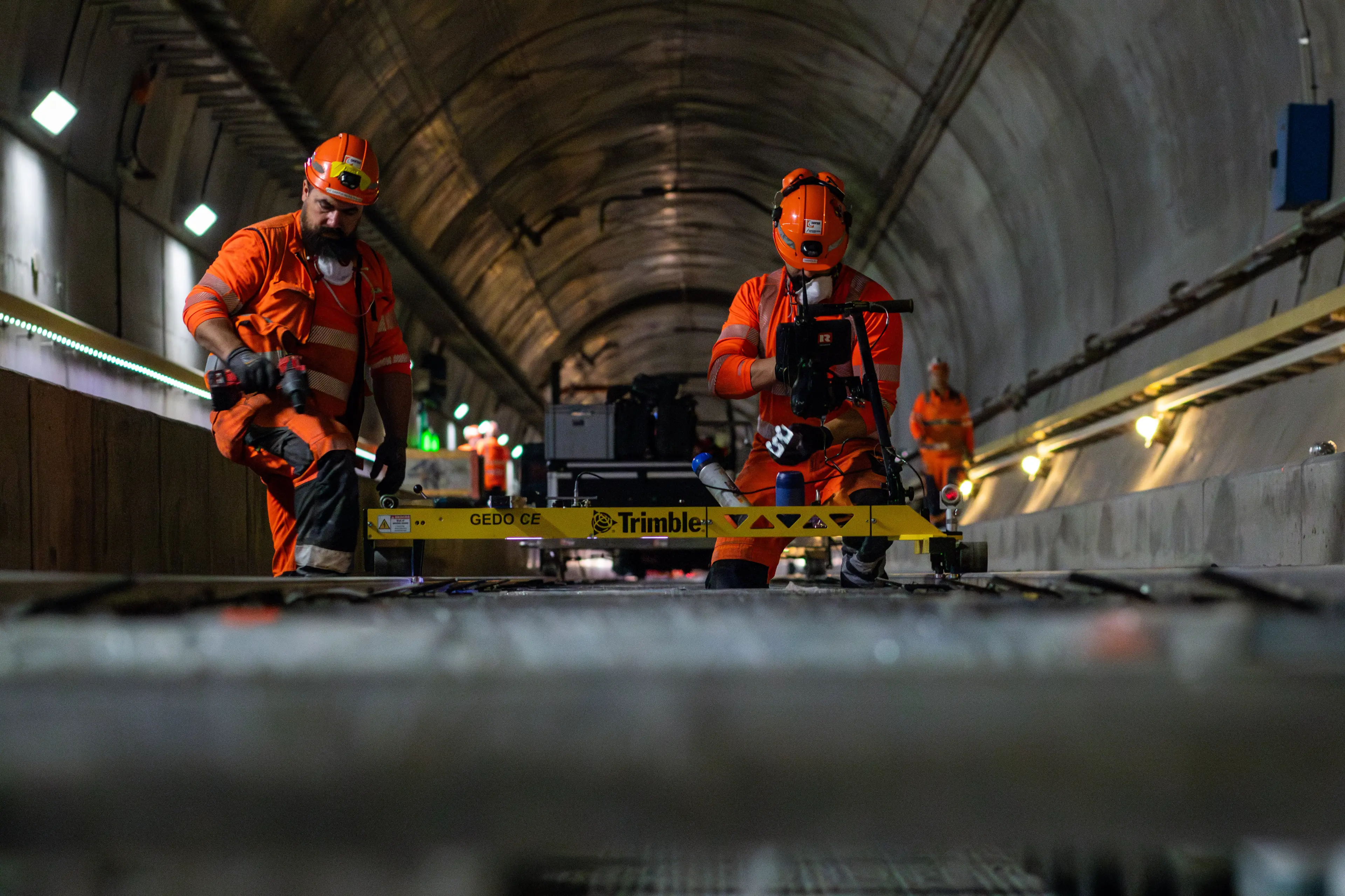 Two workers in orange safety gear use a Trimble GEDO CE measuring device on railway tracks inside a tunnel.