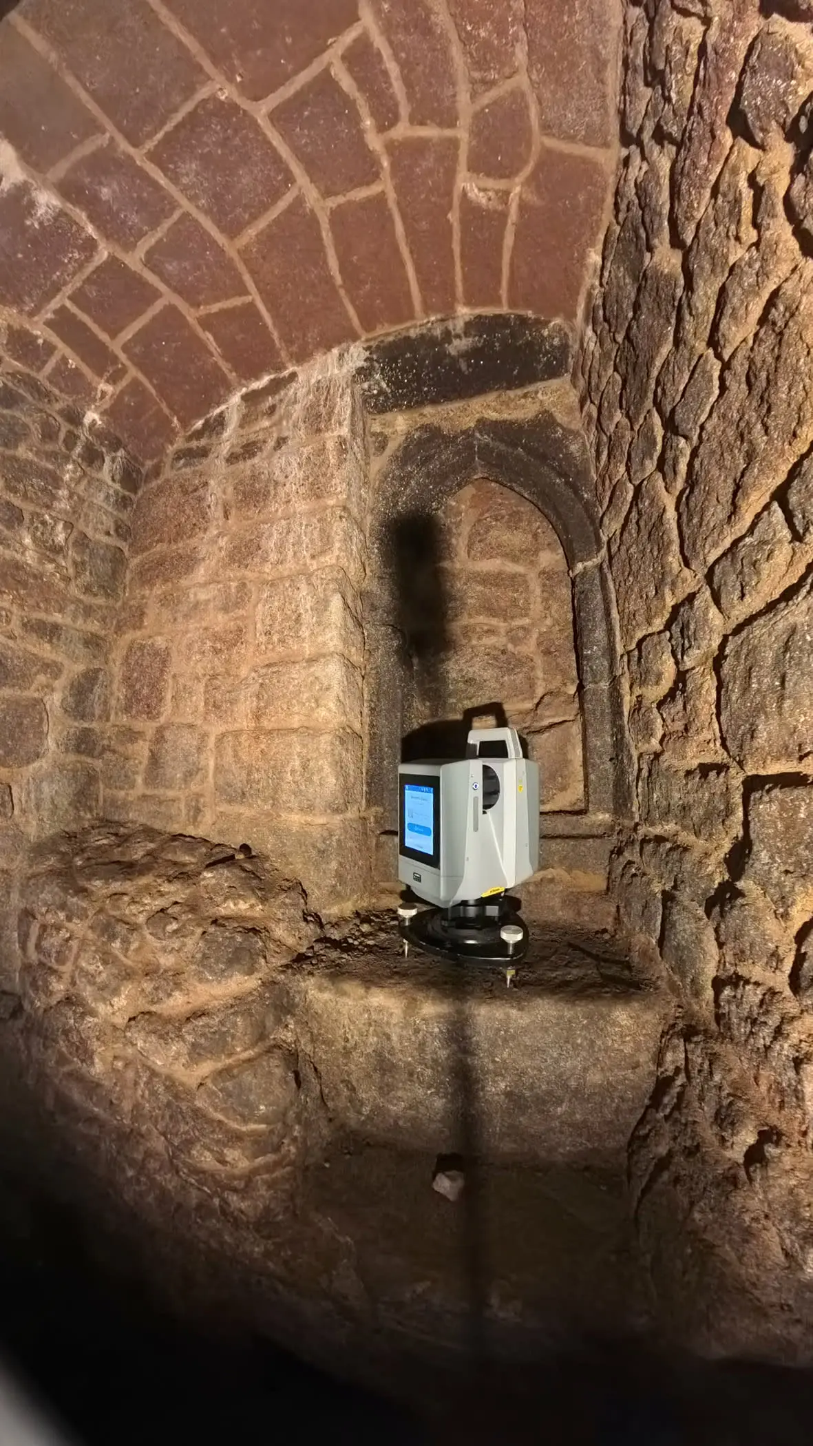 A white 3D laser scanner sits on a tripod inside an ancient stone tunnel with brick arched ceiling and alcoves.